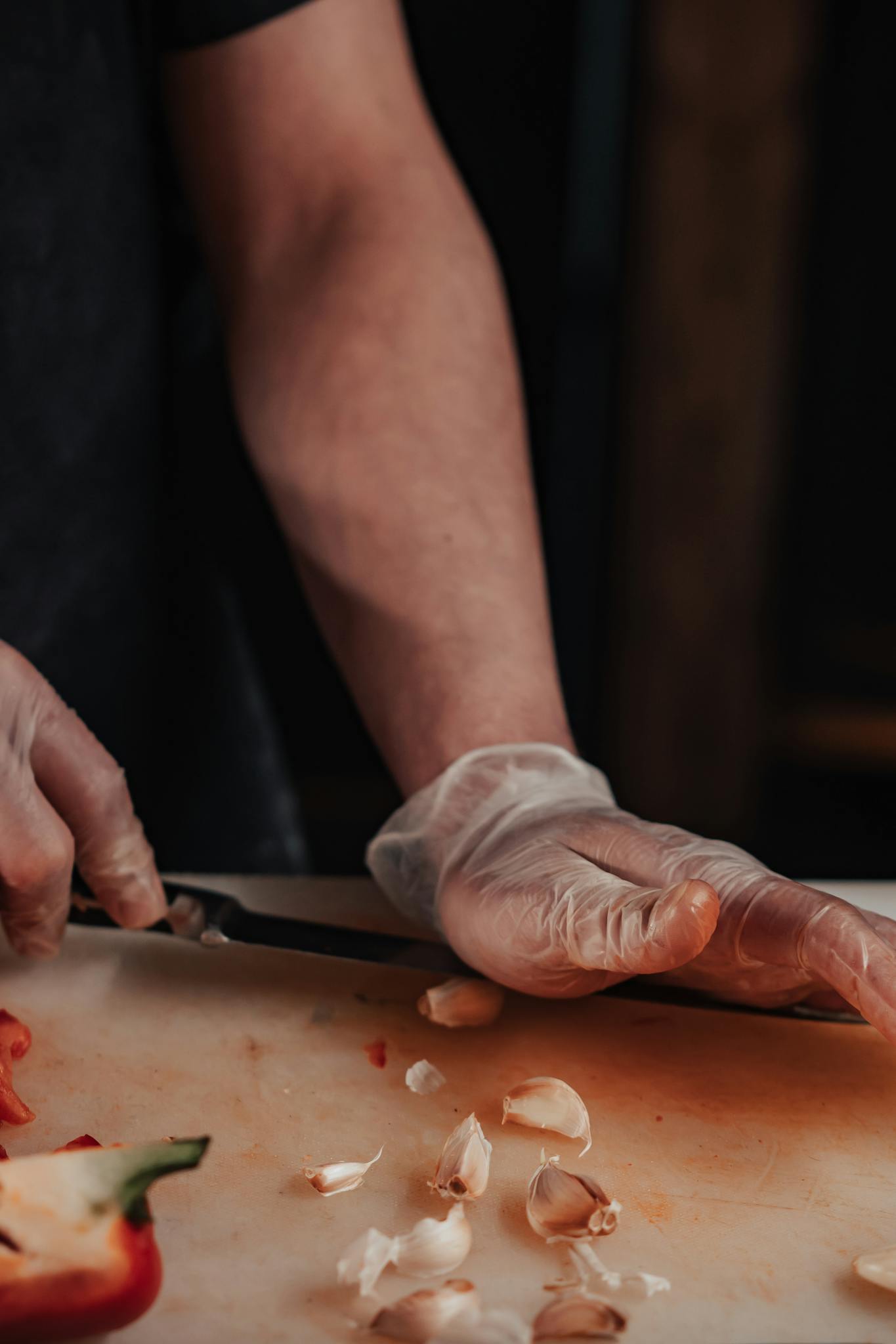 Close-up of a chef's hands wearing gloves, chopping garlic on a wooden board in a kitchen setting.