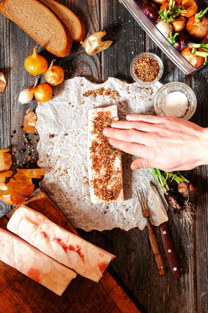 Hand seasoning pork fat with spices, surrounded by fresh ingredients on a rustic kitchen table.