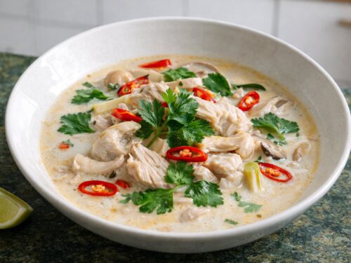 Keto Tom Kha Gai coconut chicken soup with galangal, mushrooms, fresh coriander and red chilli in a white bowl on green stone countertop